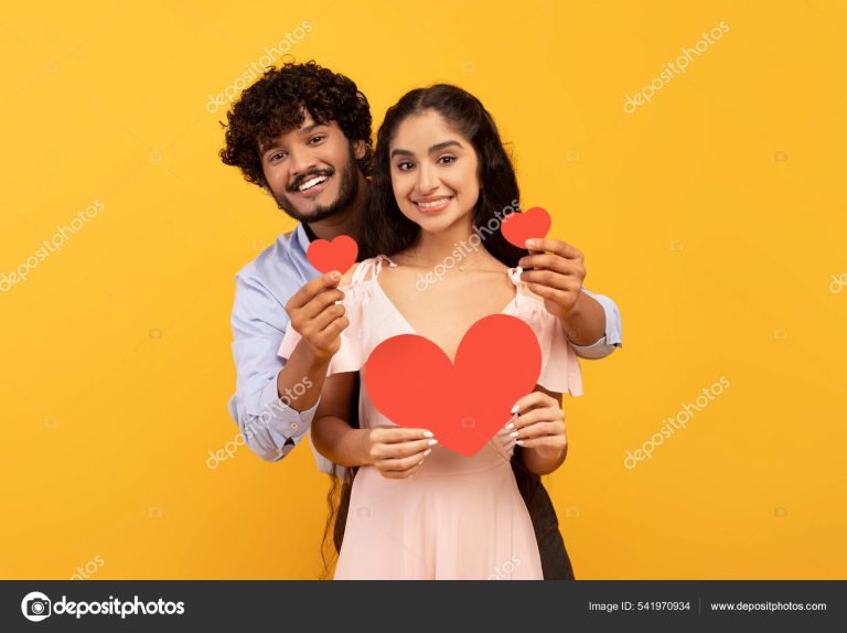 Love is in the air. Romantic indian couple with red paper hearts in hands posing over yellow studio background. Affectionate young man and woman smiling at camera