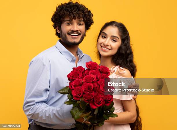 Portrait of loving indian couple with bunch of flowers smiling and looking at camera, celebrating Valentine's Day on yellow studio background. Happy spouses with roses on romantic holiday date